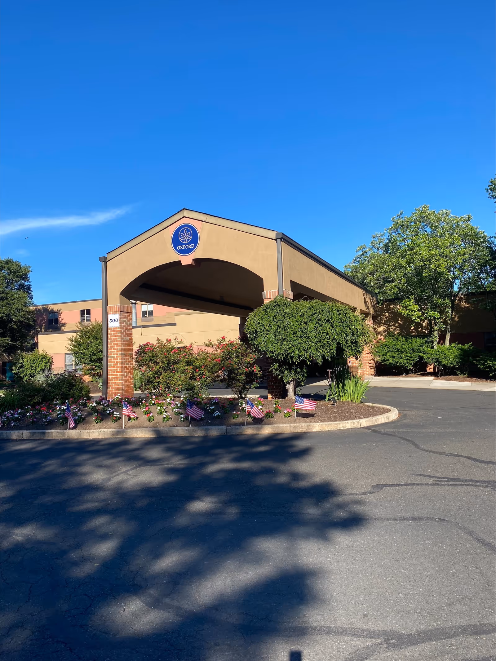 Exterior view of Oxford Rehabilitation & Healthcare Center showing the entrance canopy with the Oxford logo. The building is surrounded by greenery, including trees and flower beds with small American flags. The sky is clear and blue.