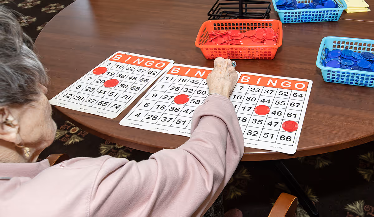 An elderly person playing bingo at a wooden table with three bingo cards and several baskets filled with red and blue bingo chips.