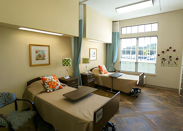 A bright and clean rehabilitation room with two hospital-style beds, each with a beige blanket and a colorful pillow. Between the beds are two nightstands with green patterned lamps. The room has large windows letting in natural light, light-colored walls, framed artwork, and wooden flooring.