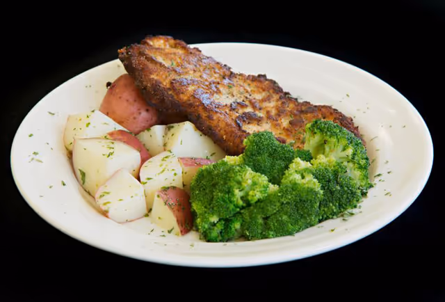 A white plate with a serving of cooked broccoli, cubed red potatoes sprinkled with herbs, and a breaded, pan-fried meat cutlet on a black background.