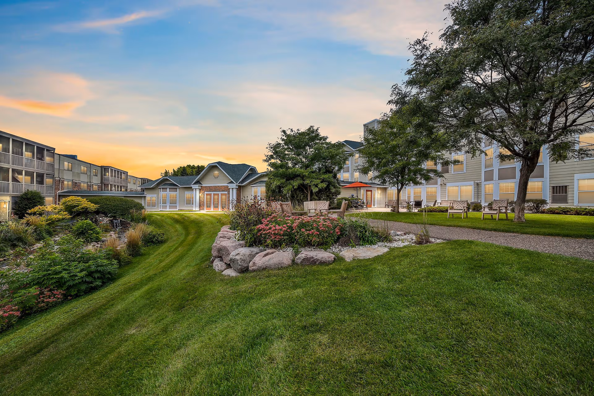 Outdoor view of a senior living facility named Waterford during sunset, showing well-maintained green lawns, landscaped garden beds with rocks and flowers, several trees, benches, and a paved walking path. The multi-story building with many windows surrounds the garden area.