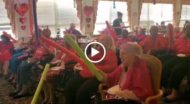 A group of elderly individuals seated in a decorated room, each holding colorful foam sticks, participating in a group activity. The room has large windows with blinds and heart-shaped decorations hanging on the walls, suggesting a festive or themed event.