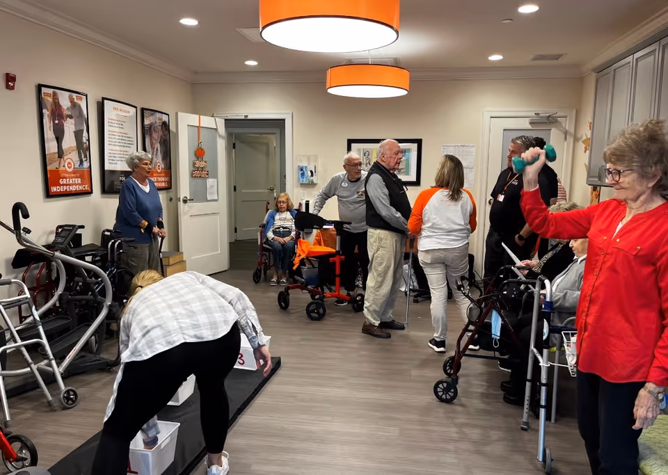 A group of elderly people and staff are gathered in a well-lit room with wood flooring and orange ceiling lights. Some seniors are using walkers and wheelchairs, while one woman in a red shirt is lifting a small green dumbbell. Another person is bending down near a game or activity on the floor. Posters are visible on the wall, and the atmosphere appears to be social and active.