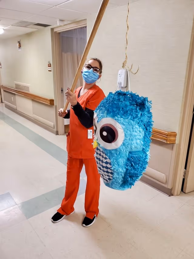 A person wearing orange scrubs and a blue face mask is standing in a hallway holding a wooden stick, about to hit a blue owl-shaped piñata hanging from the ceiling.
