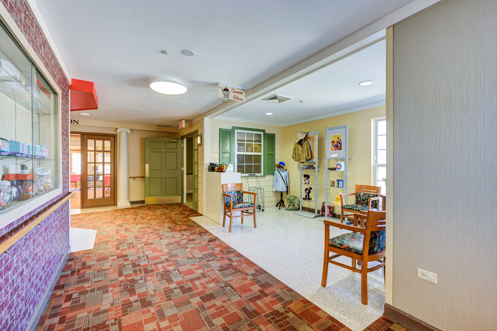 Interior view of a senior living facility hallway with a patterned carpet floor and a small corner store area featuring chairs, mannequins with clothing, and shelves with various items. There are green double doors at the end of the hallway and a glass display case on the left wall.