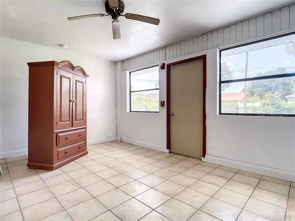 A bright, empty room with tiled floor, white walls, two large windows, a beige door with a dark red frame, a ceiling fan, and a wooden armoire.