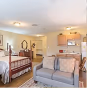 Interior view of a senior living facility room at The Manor on Lawndale featuring a bed with a wooden headboard, a dresser with a mirror, a gray sofa, and a small kitchen area with cabinets, microwave, and refrigerator.
