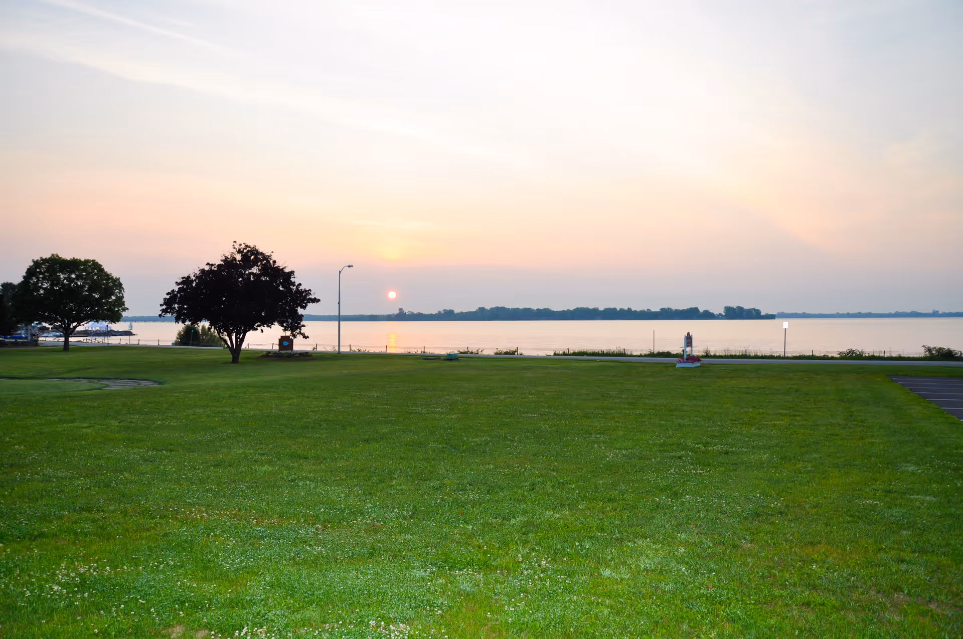 Wide grassy lawn with a few trees overlooking a calm lake at sunset.
