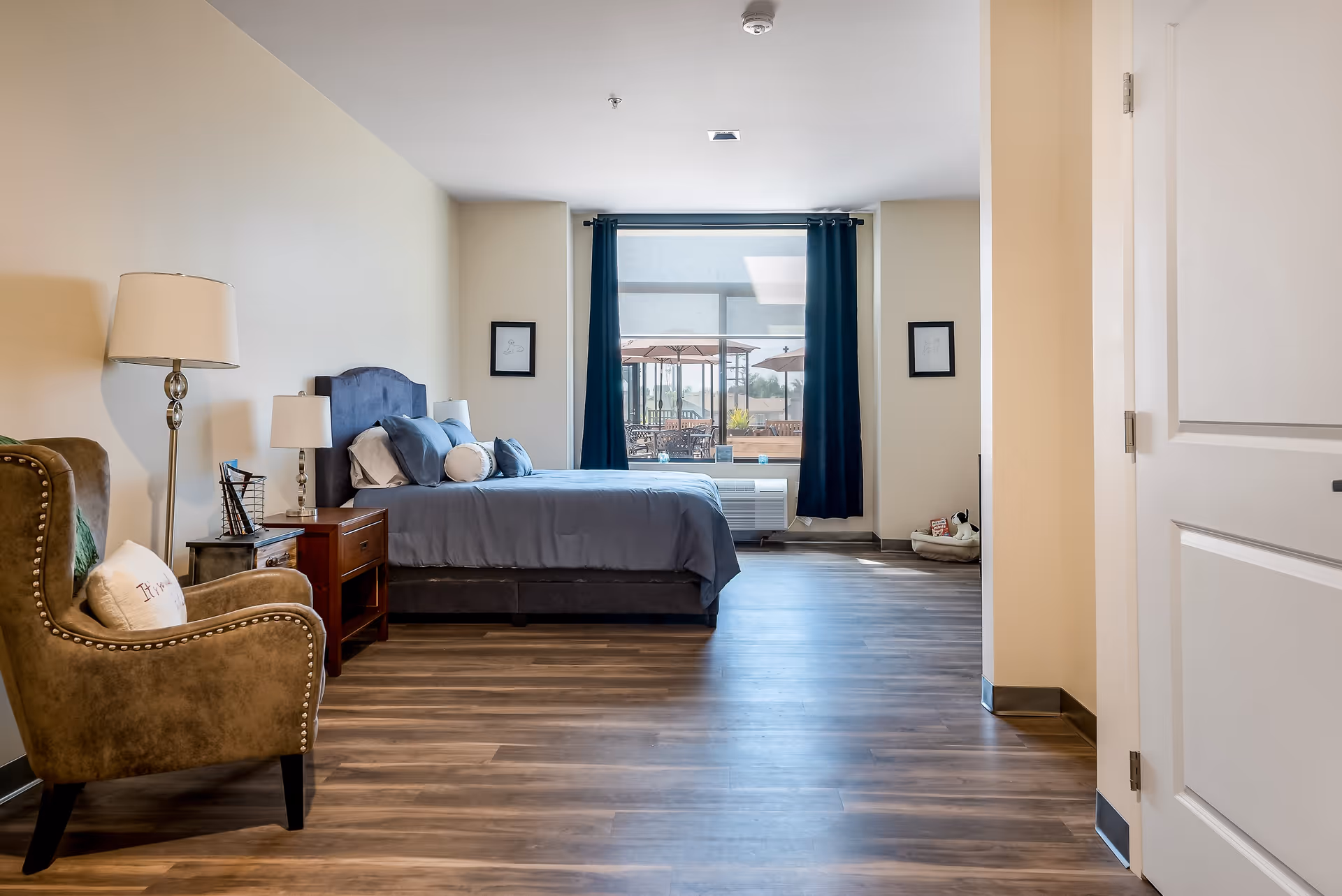 A spacious bedroom with a large window covered by dark curtains, a bed with blue bedding and multiple pillows, a wooden nightstand with a lamp, a comfortable armchair with a pillow, and a dog bed with toys in the corner near the window.