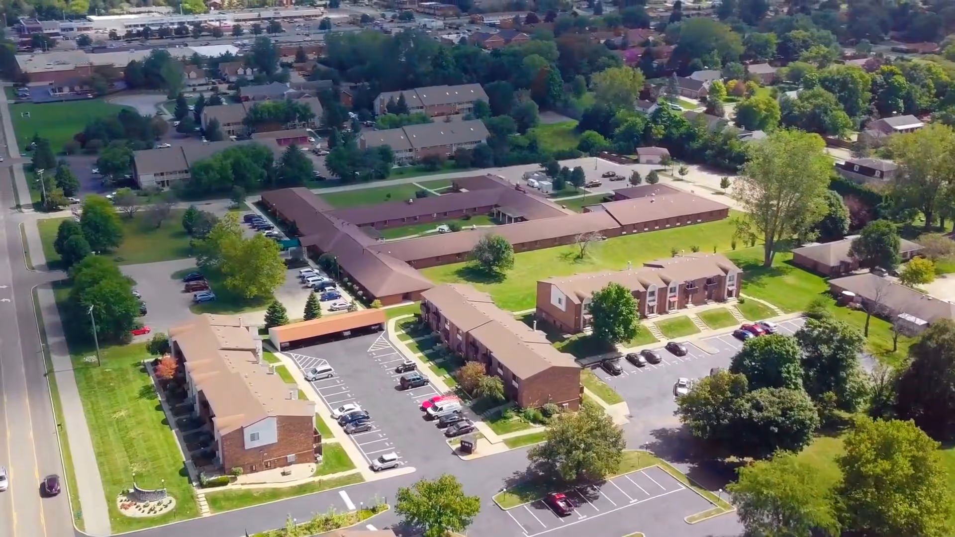 Aerial view of a senior living campus with multiple brick buildings, parking lots, roads, and surrounding trees and lawns.