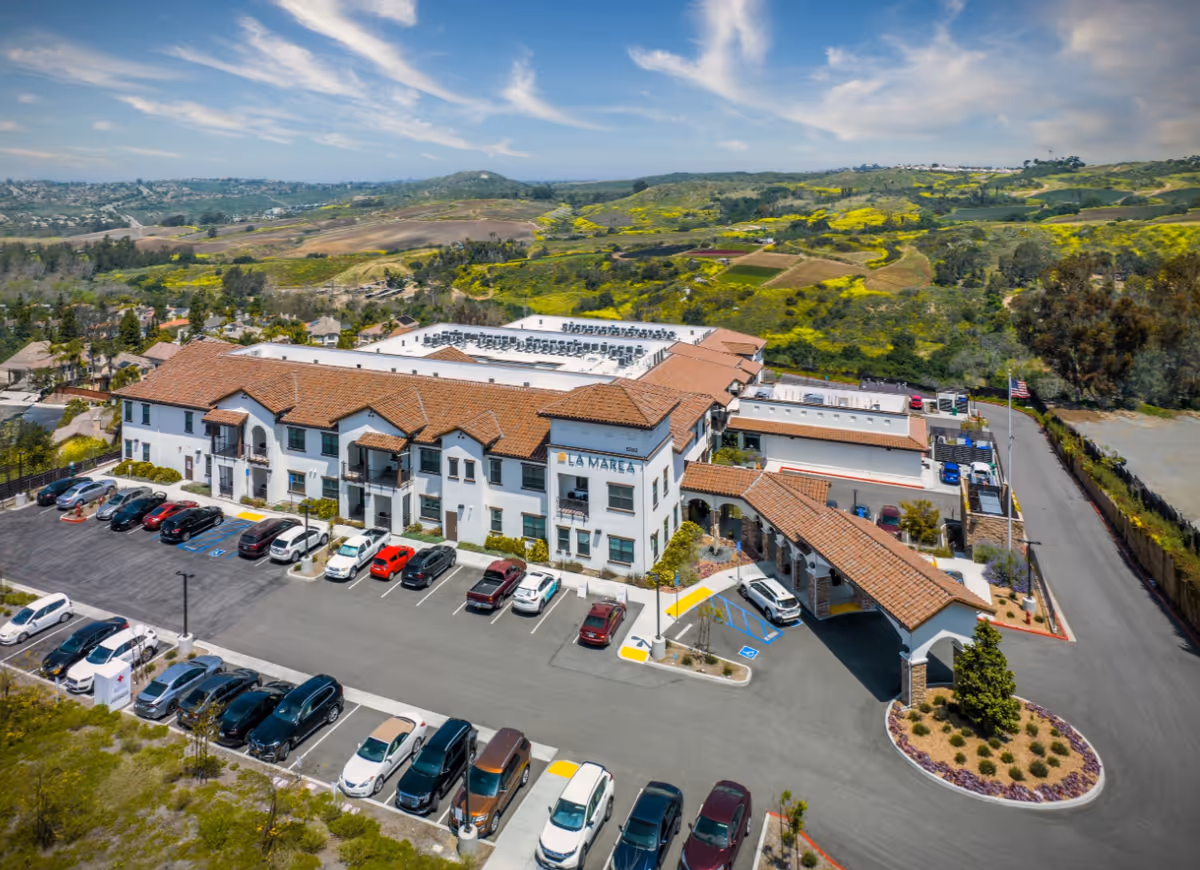 Aerial view of La Marea Senior Living's white two-story building with red tile roofs, parking lot and surrounding rolling hills.