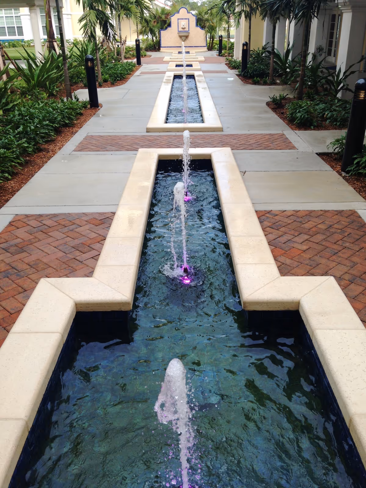 Outdoor walkway with a long rectangular water feature in the center, featuring multiple small fountains with purple lighting. The walkway is flanked by plants and bushes, with buildings visible on either side.