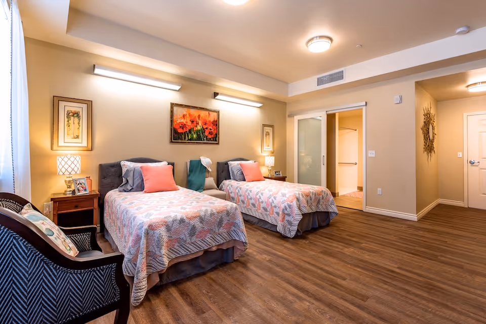 A well-lit senior living bedroom with two twin beds, each with a floral quilt and coral-colored pillow. There are two bedside tables with lamps and framed pictures, a cushioned chair with a patterned pillow, and artwork on the walls. The room has wood flooring and a sliding door leading to an adjacent room.