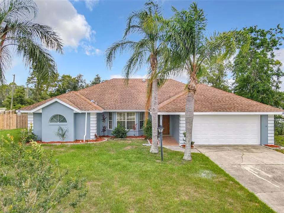 Single-story house with a brown shingle roof, light blue exterior walls, and a two-car garage. The front yard has green grass, two tall palm trees, a small lamp post, and some landscaping with plants and mulch. The sky is partly cloudy with blue patches.