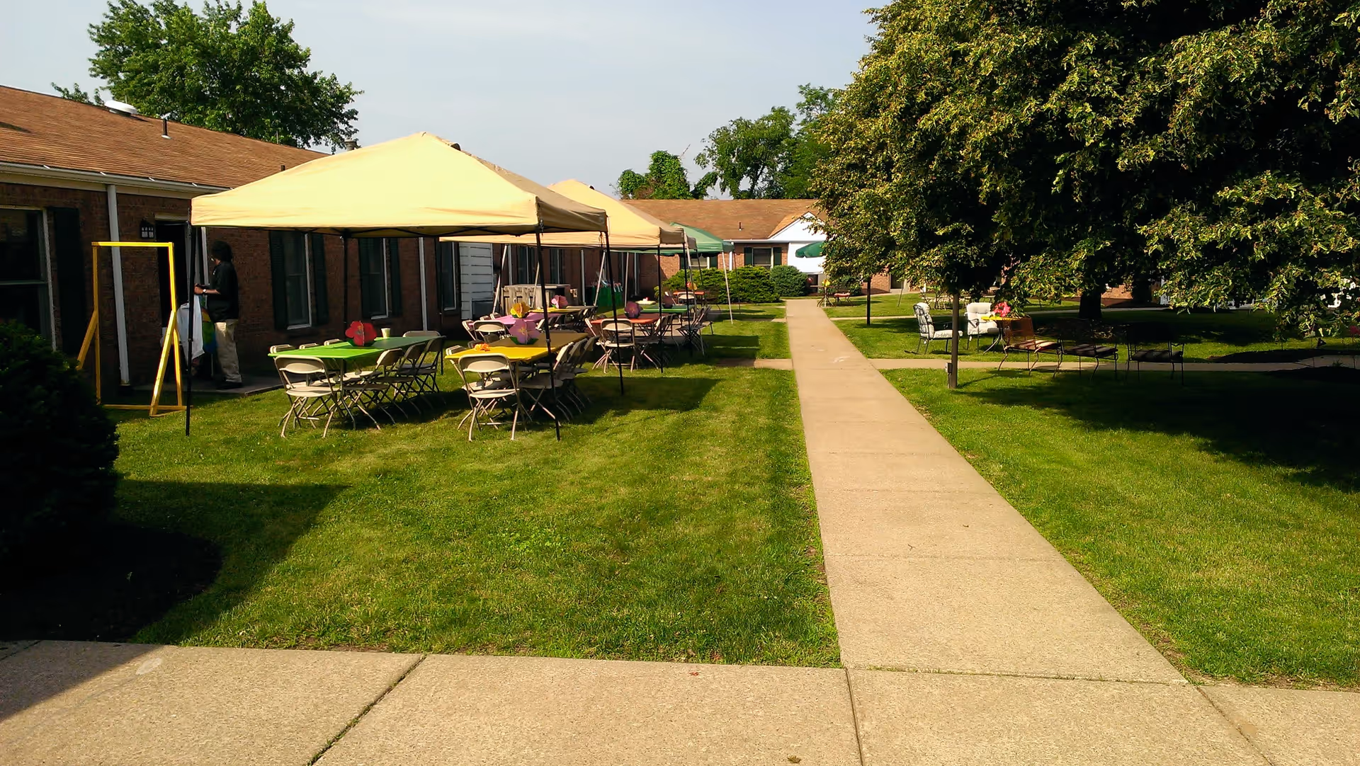 Outdoor courtyard area with a paved walkway running through the middle, surrounded by green grass and trees. On the left side, there are several tables with colorful tablecloths and folding chairs under beige canopy tents. A person is standing near the building on the left side. On the right side, there are benches and chairs under a large tree providing shade.