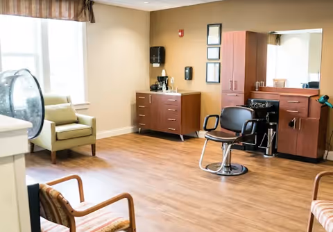 A well-lit room with wooden flooring featuring a salon chair in front of a large mirror and cabinetry. There are two armchairs with cushions near the window, and a coffee station on a cabinet against the wall.