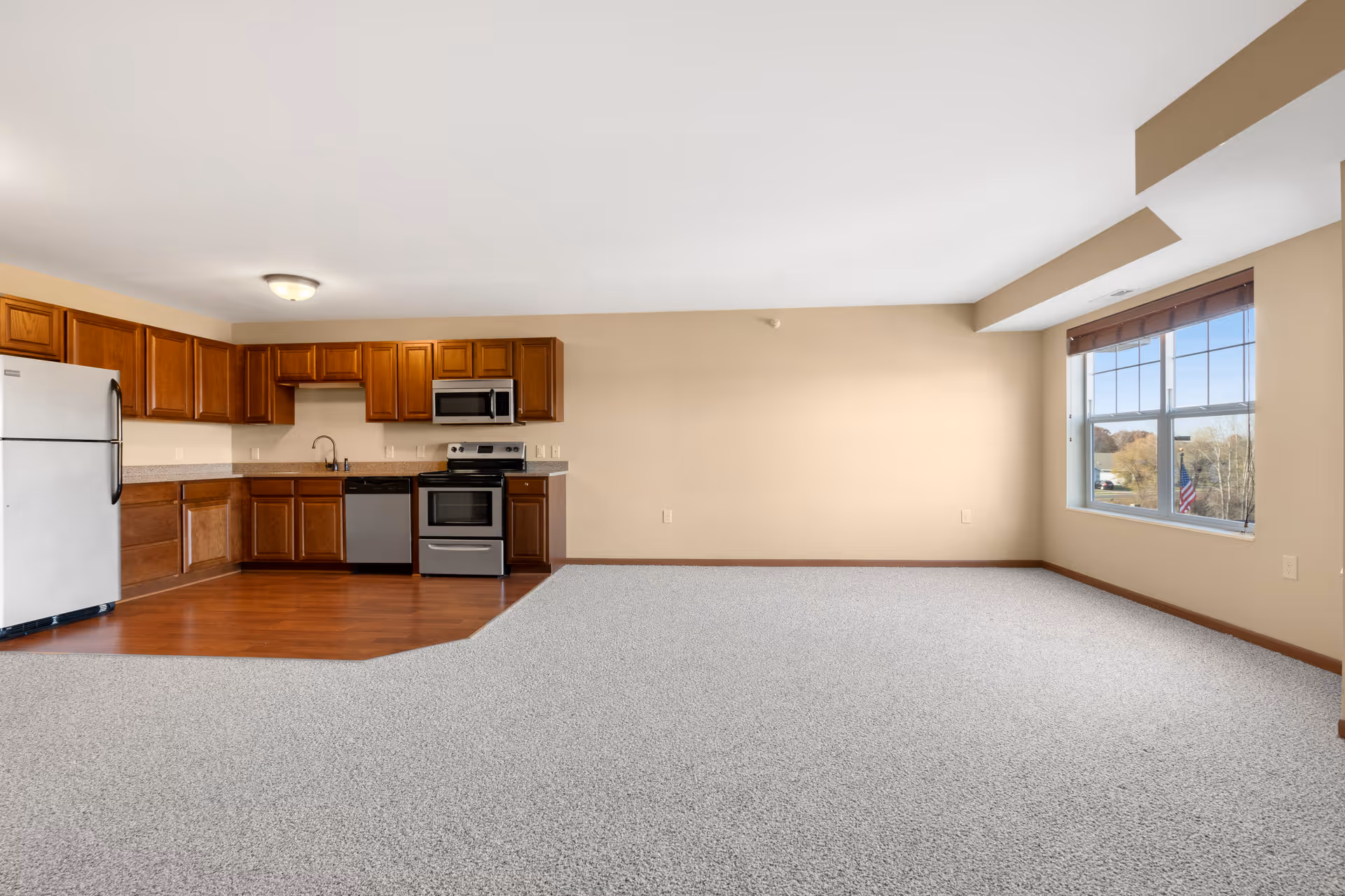 An empty open-concept living space in a senior living facility featuring a kitchen area with wooden cabinets, stainless steel refrigerator, stove, microwave, and dishwasher. The kitchen has hardwood flooring, while the adjacent living area is carpeted. A large window with blinds lets in natural light and offers a view outside.