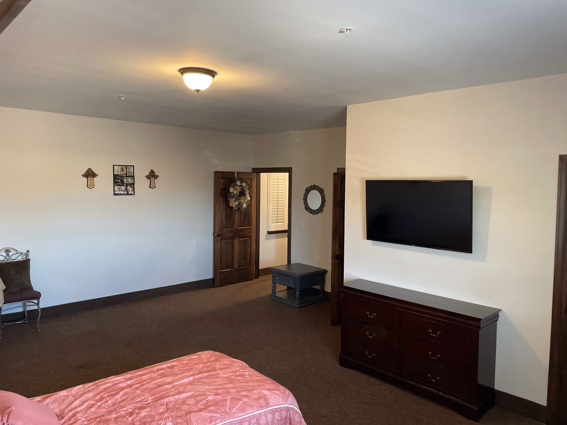 Spacious bedroom with a bed in the foreground, a wall-mounted TV above a dresser, wooden doors and simple wall decor.
