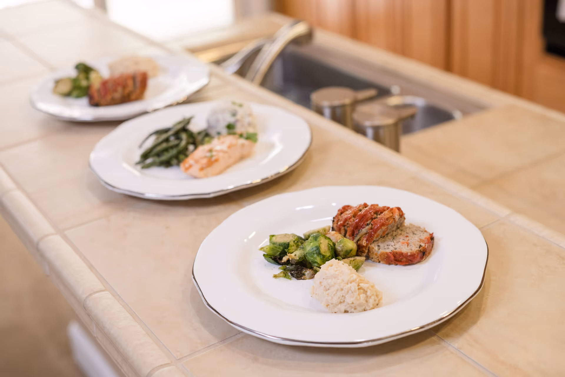 Three plated meals with vegetables and meat arranged on a tiled kitchen counter near a sink.