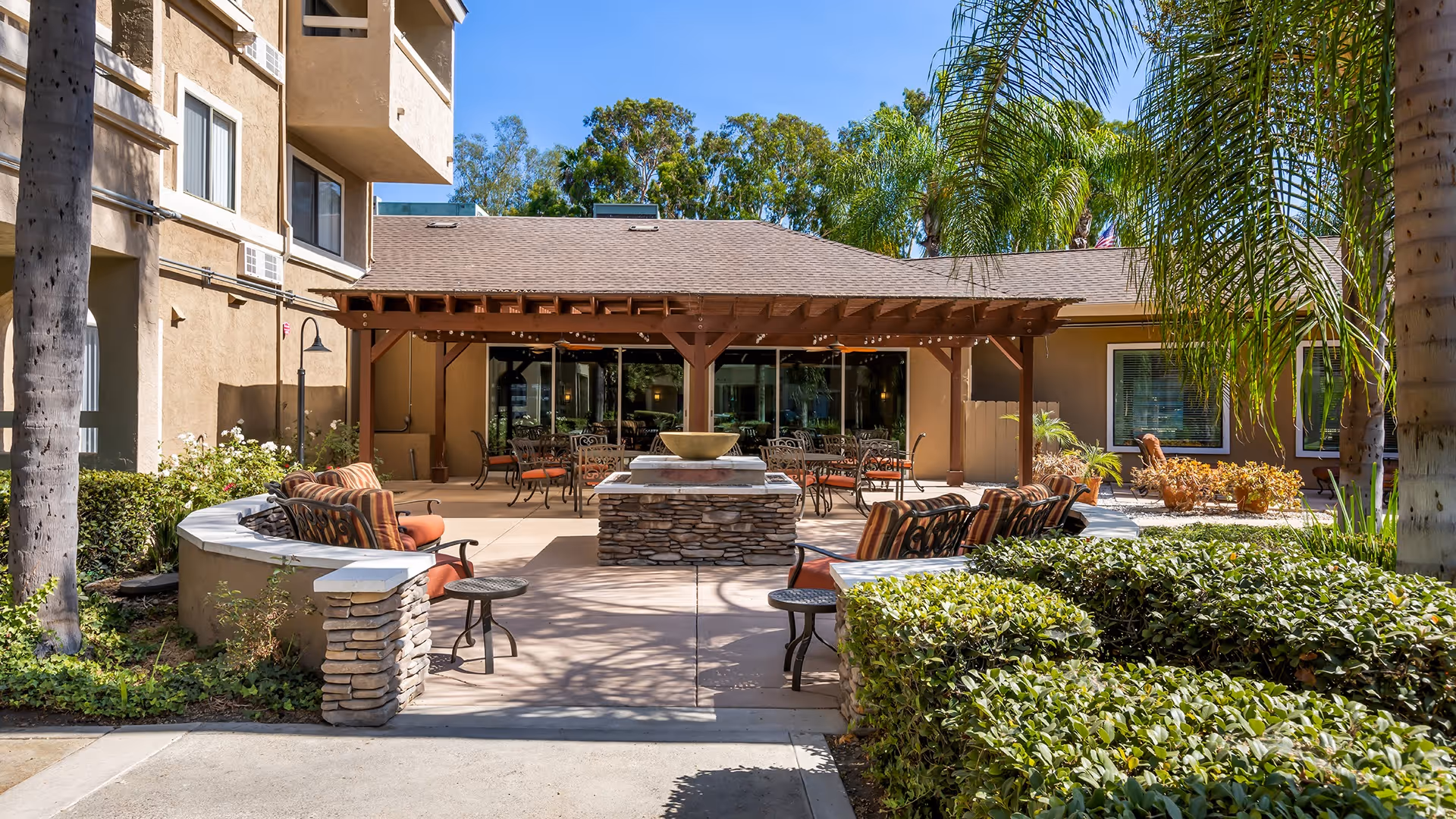 Outdoor patio area at Holiday Springs of Escondido featuring cushioned seating around a central stone fire pit, a pergola with string lights, and surrounding greenery including palm trees and bushes.