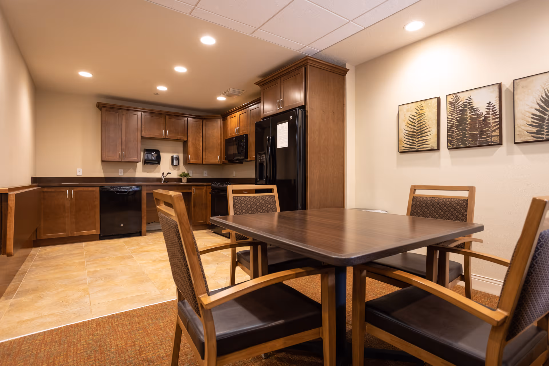 A kitchen and dining area with wooden cabinets, black appliances including a refrigerator, dishwasher, and microwave. There is a wooden dining table with four chairs in the foreground, and three framed botanical prints on the wall. The room has recessed ceiling lights and a tiled floor transitioning to carpet near the dining area.
