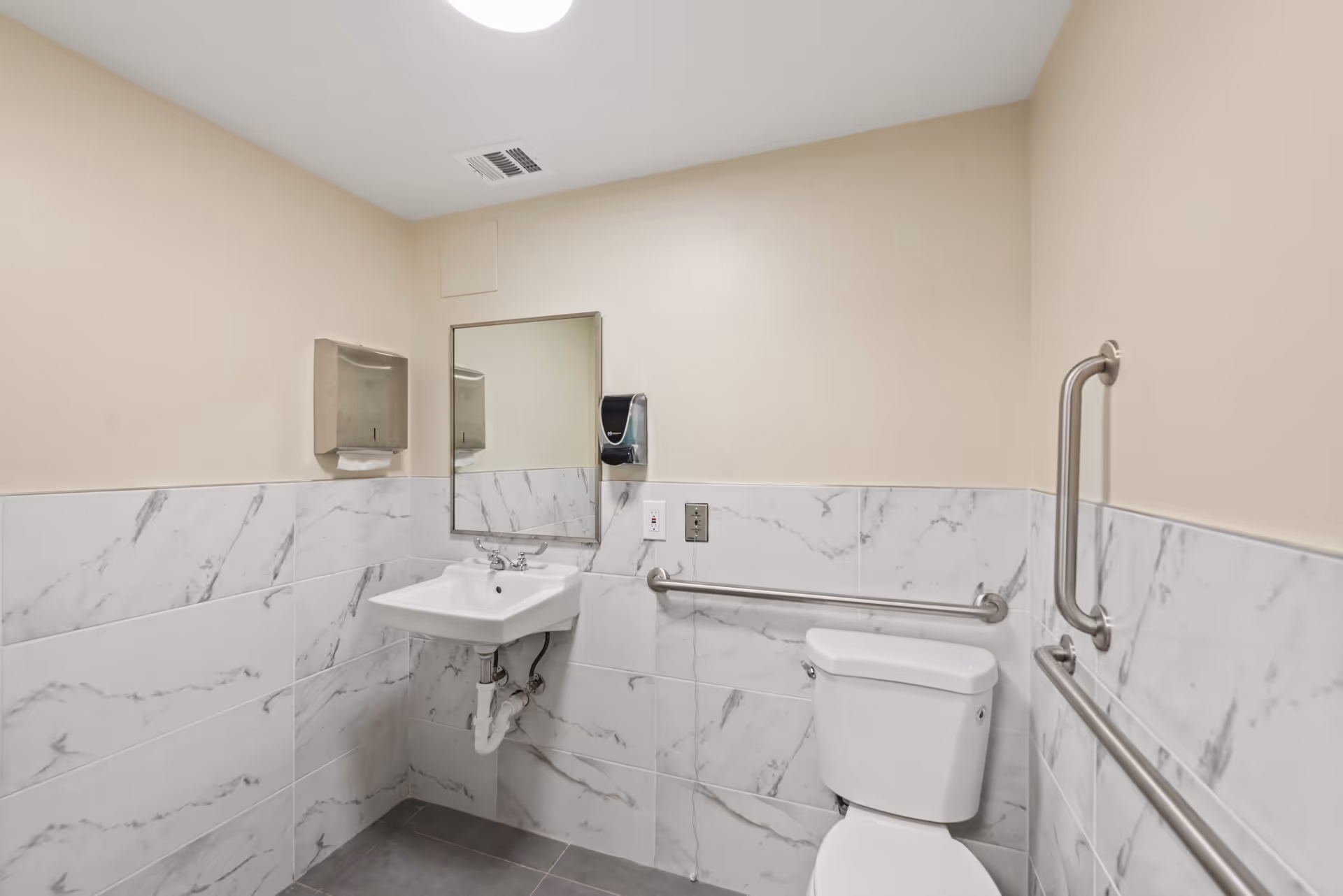 A clean and accessible bathroom with beige walls and marble-patterned tiles halfway up. It features a white toilet with stainless steel grab bars on both sides, a white wall-mounted sink with exposed plumbing, a rectangular mirror above the sink, a wall-mounted soap dispenser, and a paper towel dispenser.