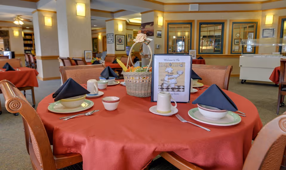 A dining table set for four with a red tablecloth, black folded napkins, bowls, plates, cups, and silverware. A basket with decorative items and a framed sign that reads 'Welcome to The Bistro' featuring a chef cartoon are placed in the center. The background shows more tables with similar settings and warm lighting fixtures on the walls.