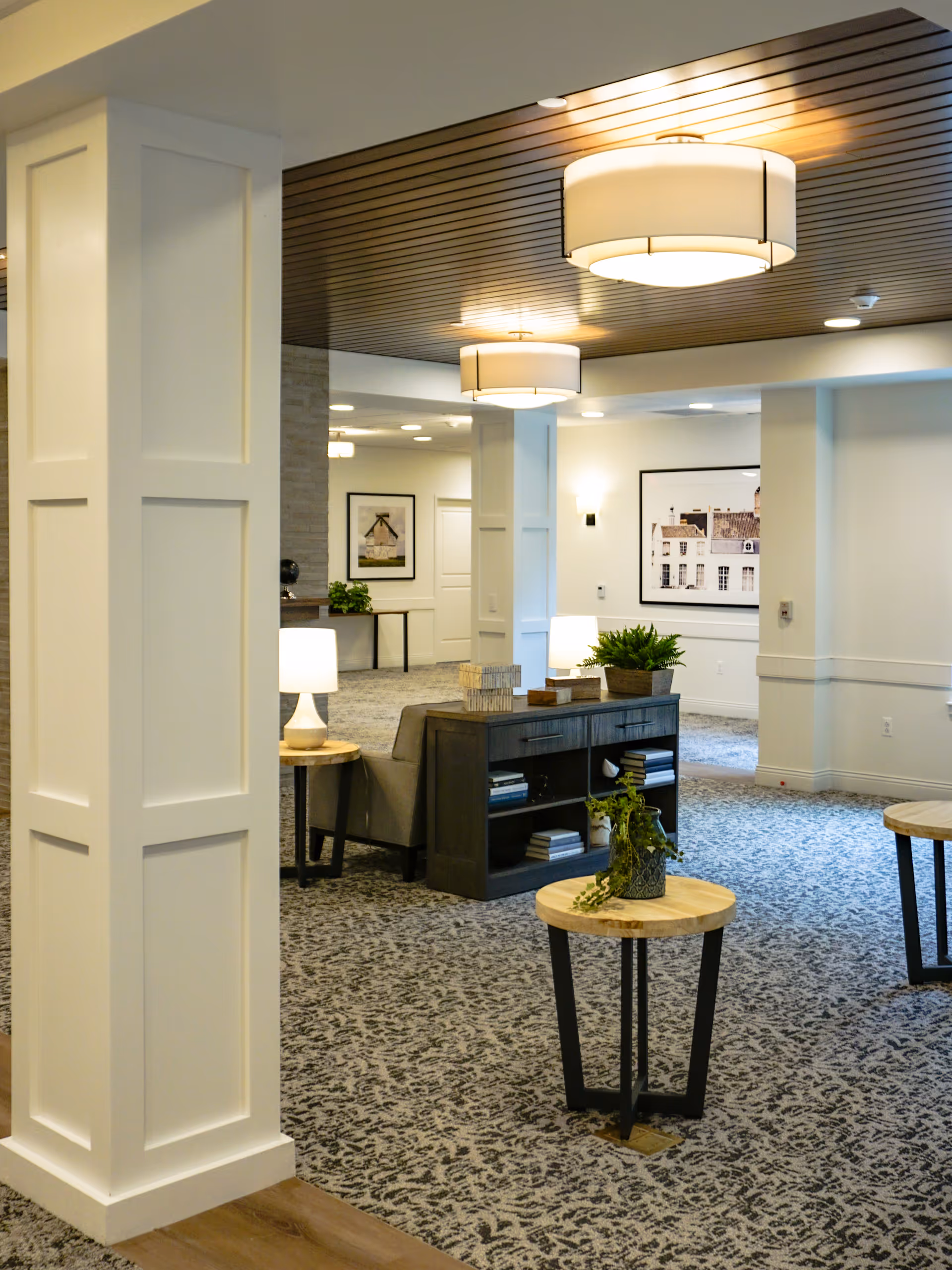 Bright modern senior living lobby with seating, side tables, plants, framed artwork, and hanging ceiling lights.