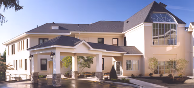 Exterior view of a two-story senior living facility building with a covered entrance supported by stone pillars, large windows, and a well-maintained driveway and landscaping under a clear blue sky.