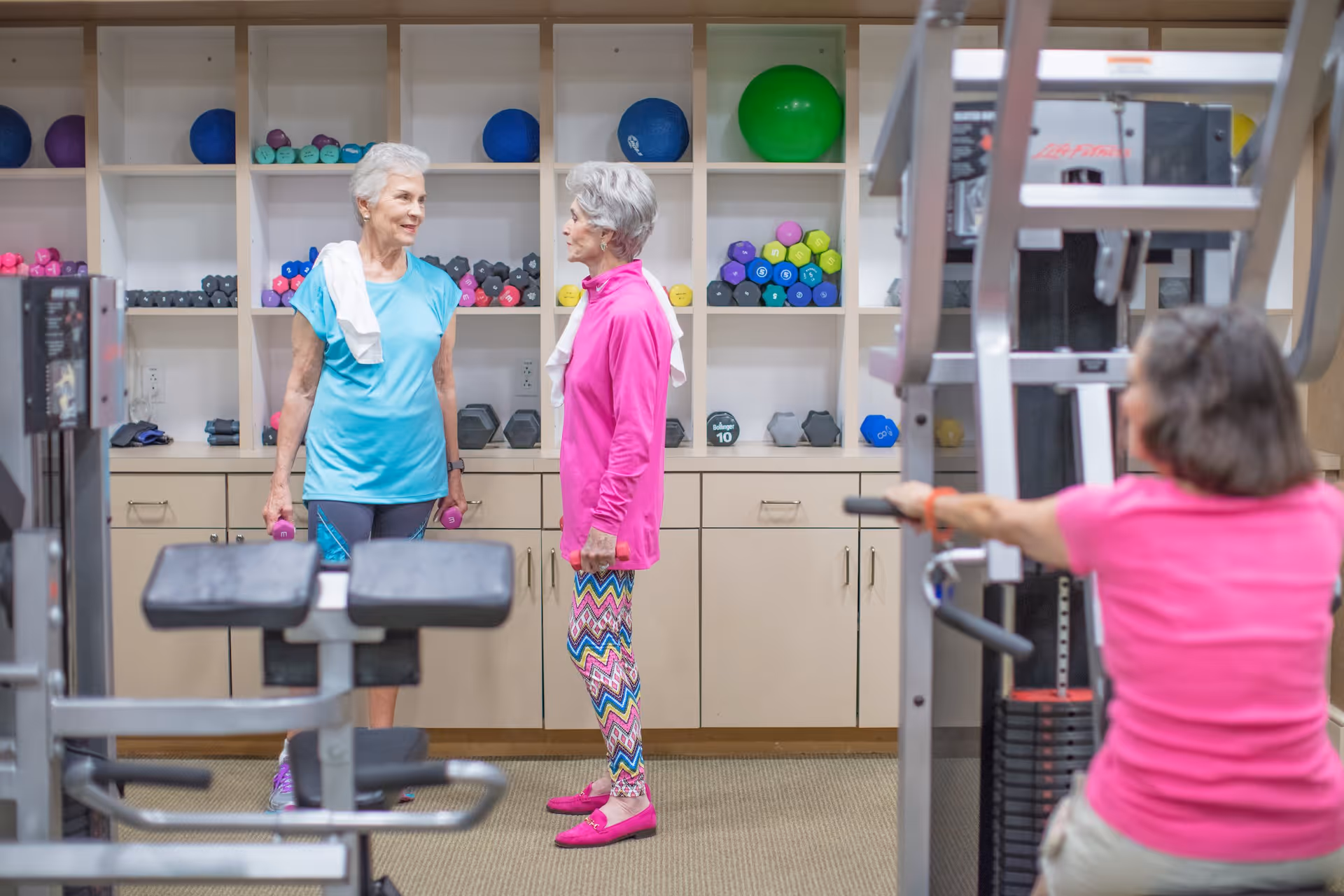 Two elderly women standing and talking in a fitness room with shelves of colorful dumbbells and exercise balls behind them. Another woman is using a weight machine in the foreground.
