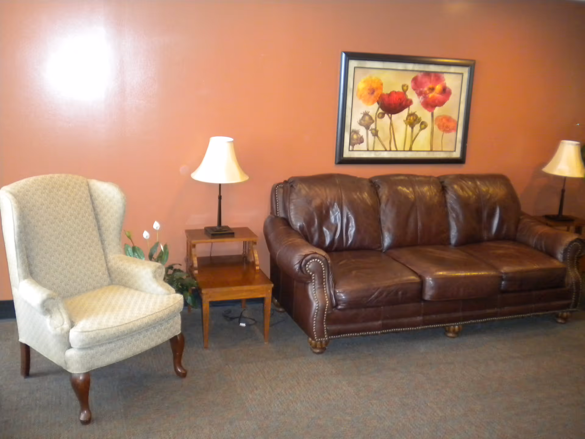 Leather sofa and upholstered armchair with side tables, lamps, and floral artwork on a rust-colored wall.
