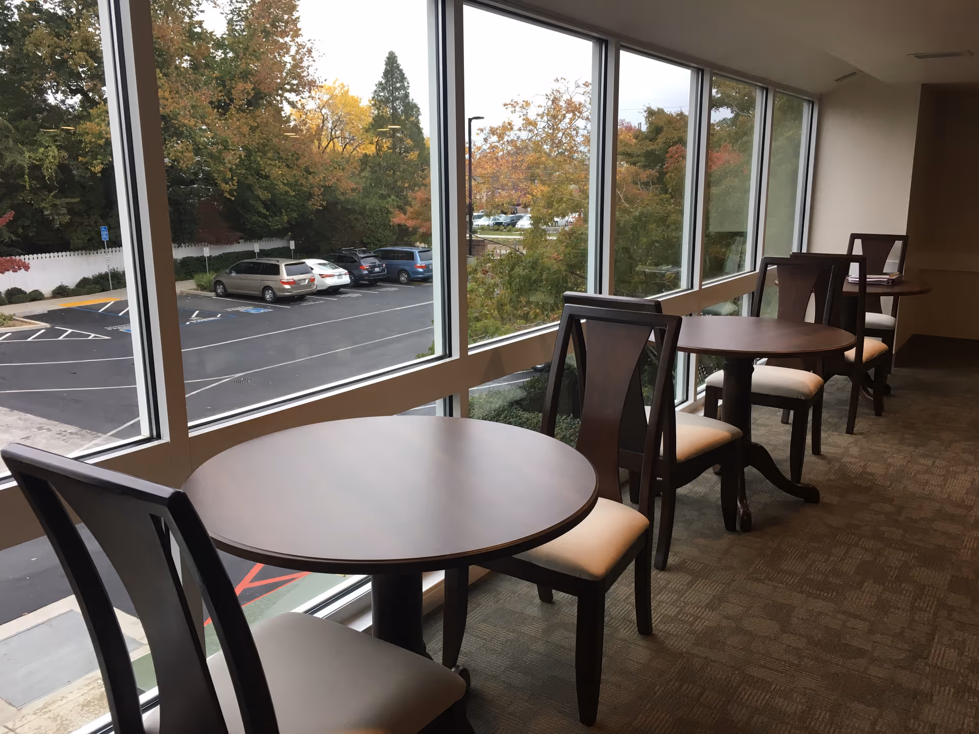 Interior view of a seating area with round wooden tables and cushioned chairs lined up along large windows overlooking a parking lot and trees with autumn foliage.