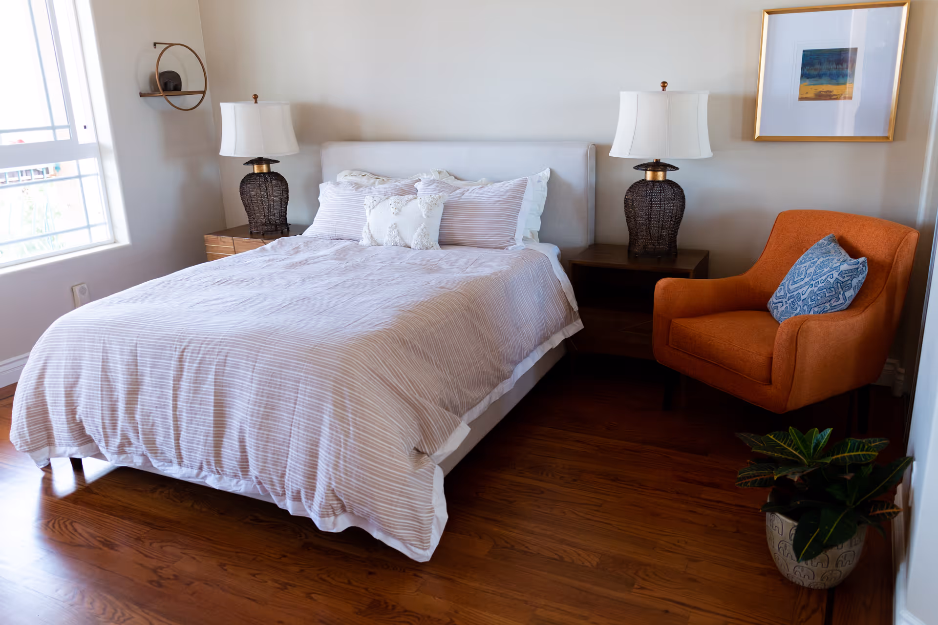 A bedroom with a neatly made bed featuring striped bedding and multiple pillows. On either side of the bed are dark wooden nightstands with matching lamps. To the right of the bed is an orange armchair with a blue patterned cushion. A framed artwork hangs on the wall above the chair, and a potted plant is placed on the floor nearby. A window on the left side allows natural light into the room.