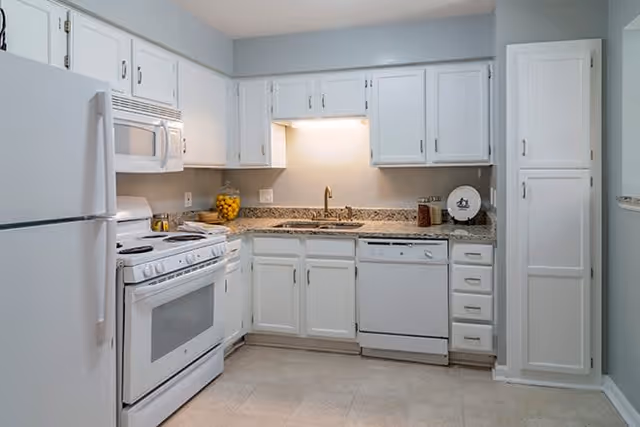 A clean and bright kitchen with white cabinets, a white refrigerator, a white stove with oven, a white microwave above the stove, a dishwasher, and a granite countertop. There is a sink under a cabinet with a light above it, and some kitchen items including a bowl of lemons and a decorative plate on the counter.