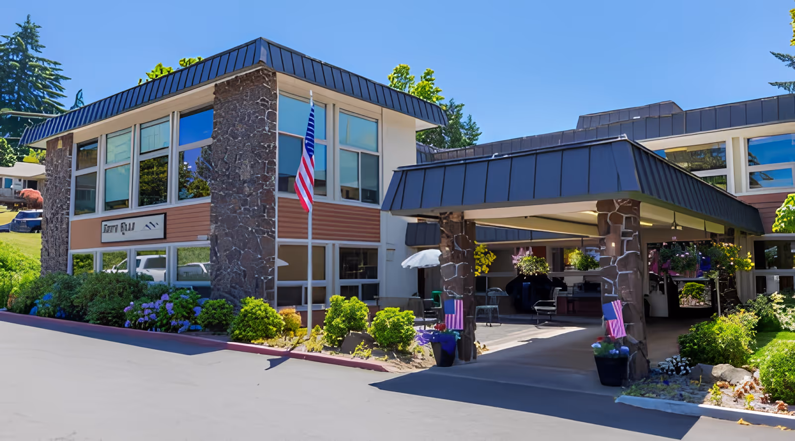 Exterior view of South Hills Rehabilitation Center building with stone and wood paneling, large windows, an American flag on a pole, and a covered entrance with hanging flower baskets and outdoor seating. The surrounding area has green bushes and trees under a clear blue sky.