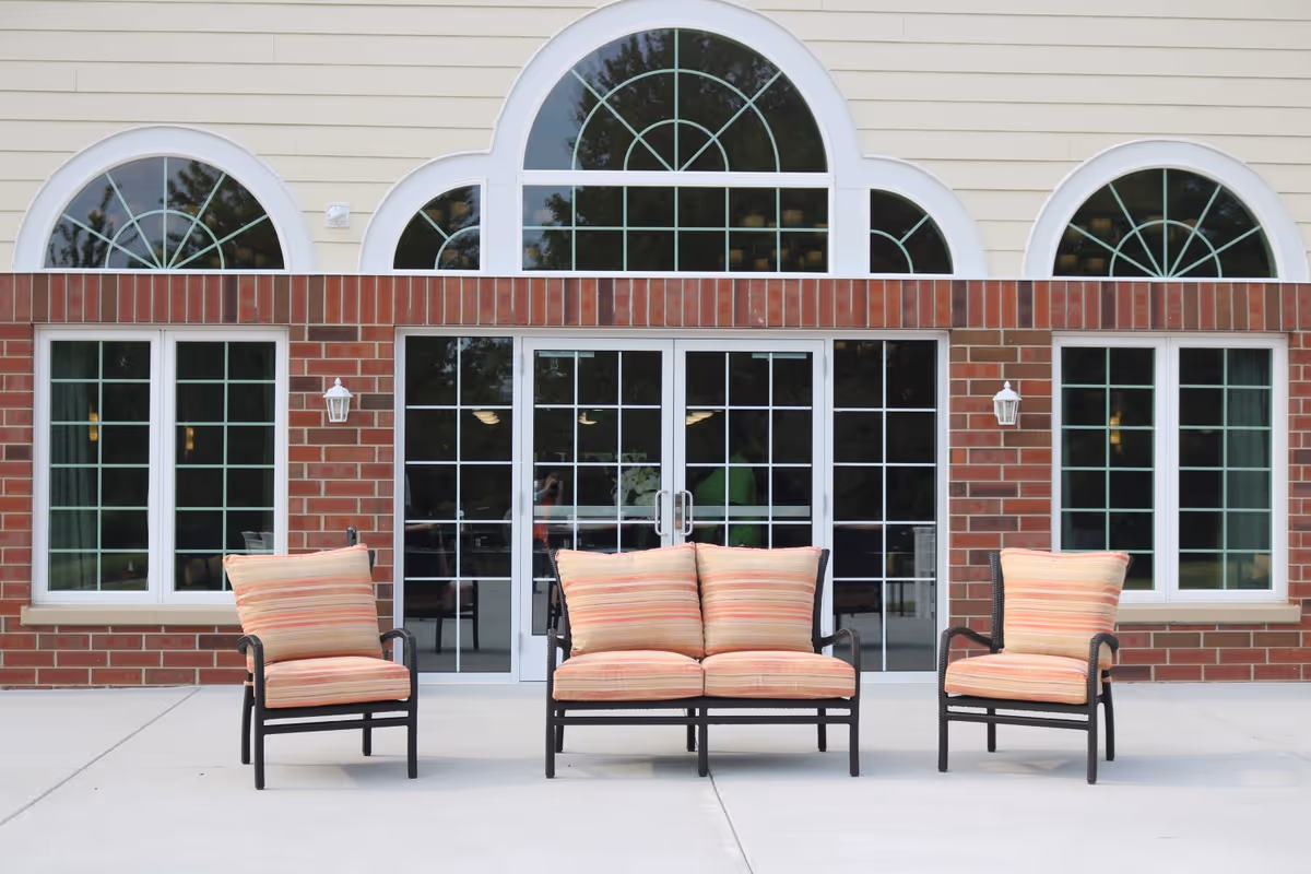 Outdoor patio area with three cushioned chairs arranged in front of a building with large windows and a brick facade.