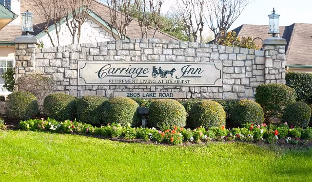 Stone entrance sign for Carriage Inn Retirement Living at its Finest located at 2805 Lake Road, surrounded by neatly trimmed bushes and colorful flowers with residential buildings in the background.