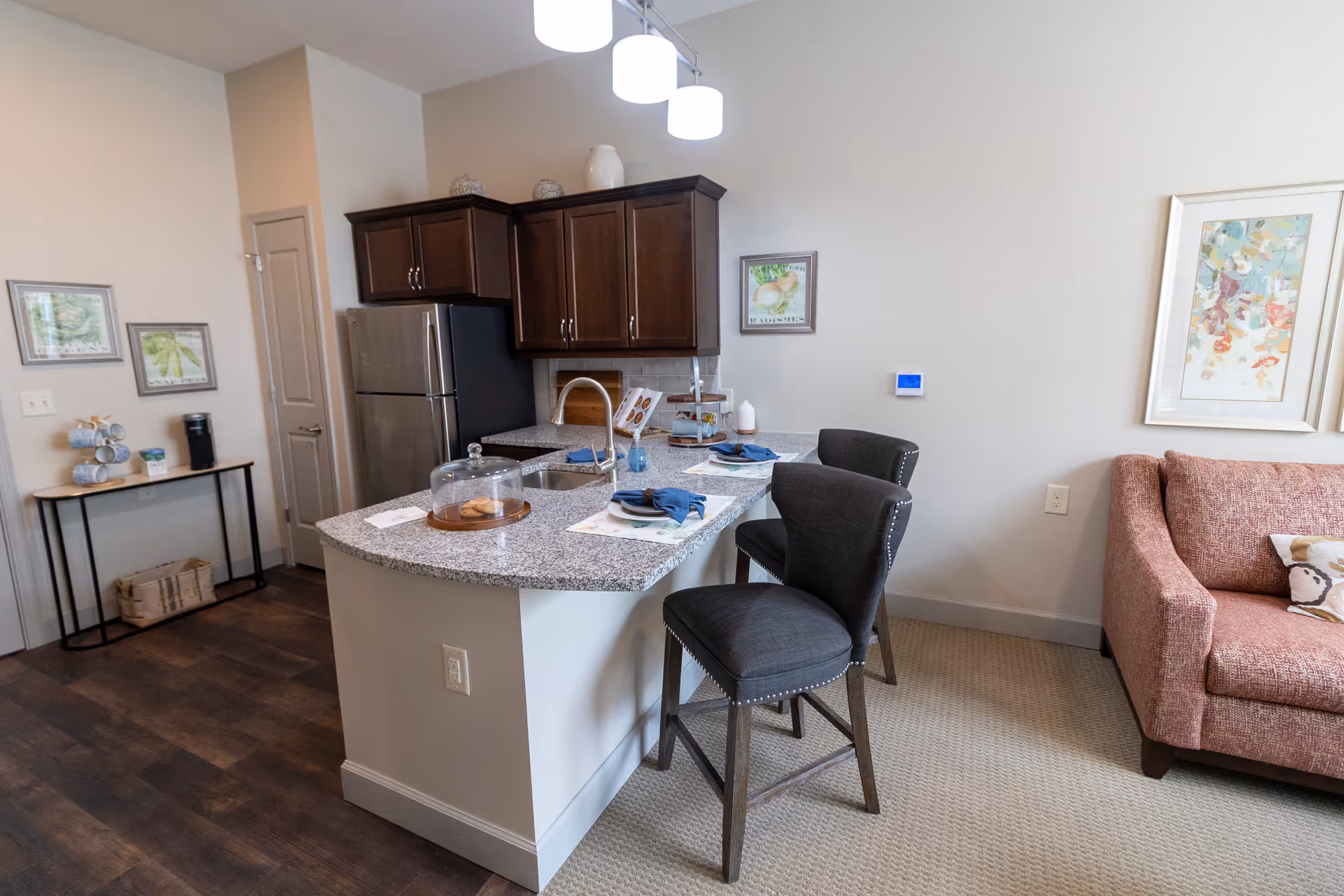 Interior view of a modern kitchen and living area in a senior living facility. The kitchen features dark wood cabinets, a stainless steel refrigerator, and a granite countertop island with a sink and two dark upholstered bar stools. On the countertop, there is a glass dome with cookies, a tiered tray, and place settings with blue napkins. The living area includes a pinkish sofa with decorative pillows and framed artwork on the walls. The floor transitions from dark wood in the kitchen area to light carpet in the living space.