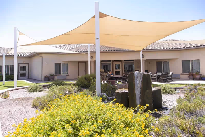 Outdoor patio area at Copper Canyon facility with yellow shade sails overhead, a stone water fountain surrounded by greenery and yellow flowers, and several tables and chairs arranged for seating near the building.