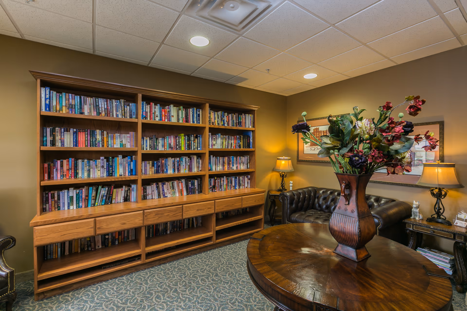 A cozy interior room with a large wooden bookshelf filled with books, a round wooden table with a decorative vase of flowers, a dark leather tufted sofa, two side tables with lamps, and framed artwork on the wall.