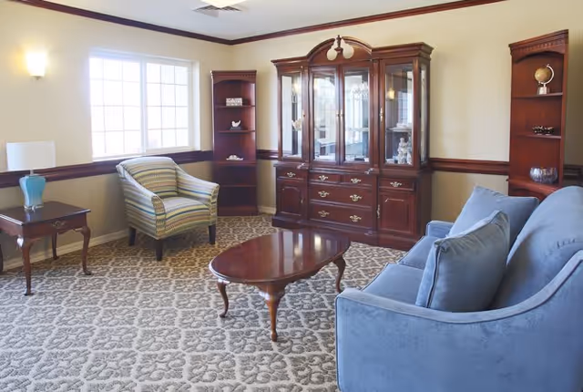 A cozy living room in an assisted living facility featuring a blue upholstered sofa with cushions, a wooden coffee table, a striped armchair, a wooden side table with a blue vase, and a large wooden china cabinet with glass doors. The room has patterned carpet flooring, cream-colored walls with a wooden chair rail, and a large window letting in natural light.