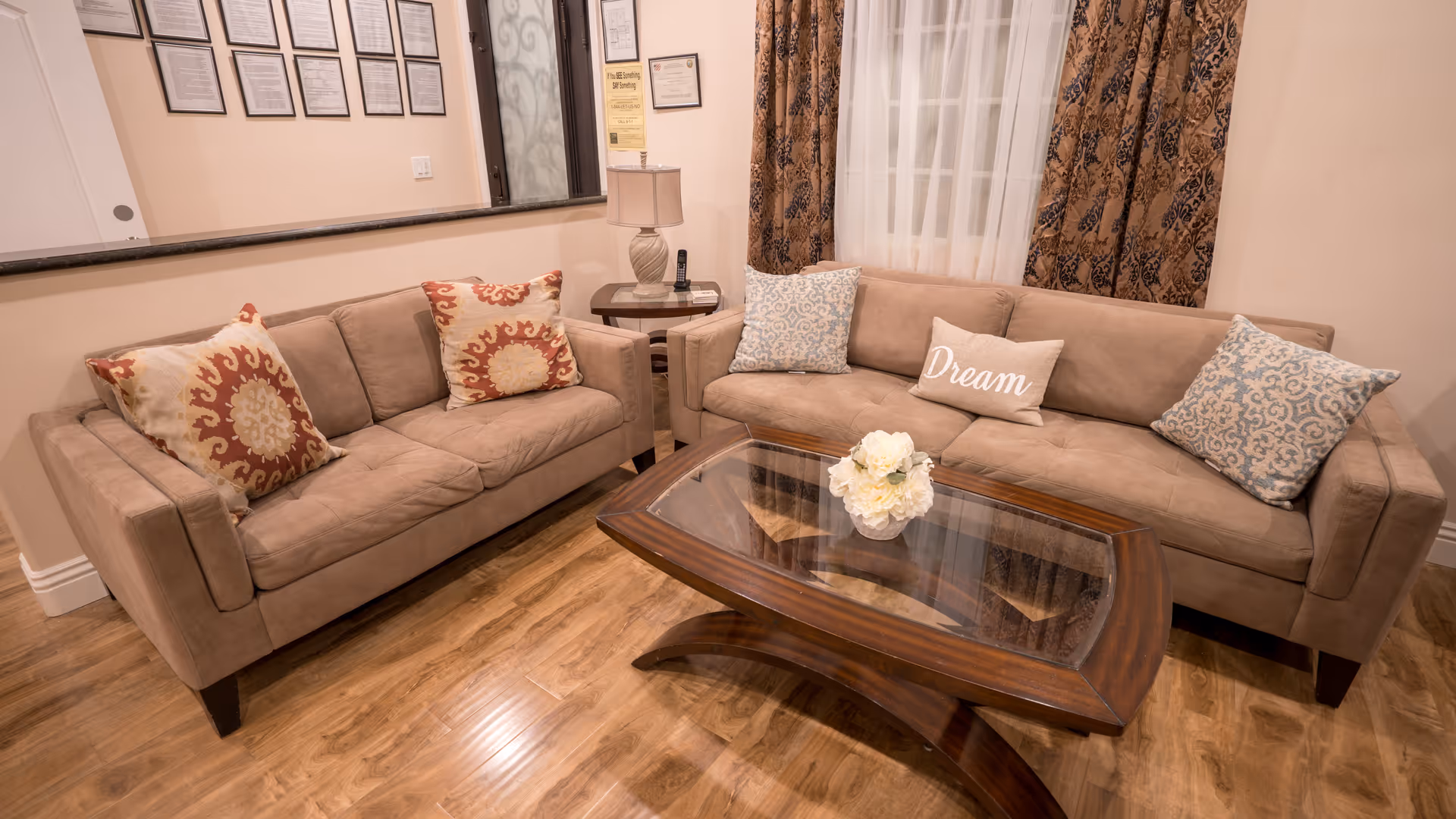A cozy living room with two beige sofas adorned with decorative pillows, including one with the word 'Dream'. A wooden coffee table with a glass top and a small flower arrangement sits in front of the sofas. The room has wooden flooring, patterned curtains, a side table with a lamp and a phone, and framed documents on the wall.
