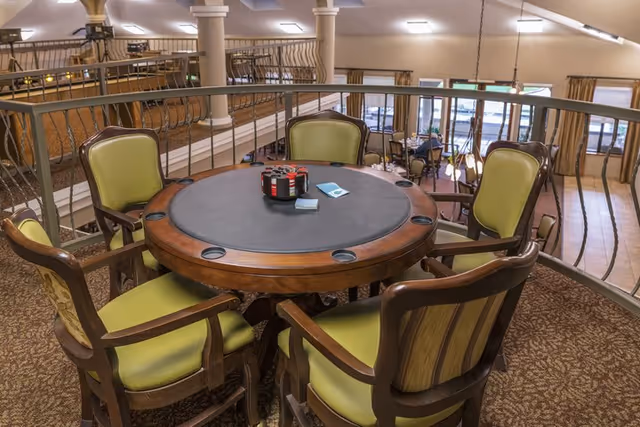 Round card table with poker chips and four green-upholstered chairs on an indoor balcony overlooking a dining area.