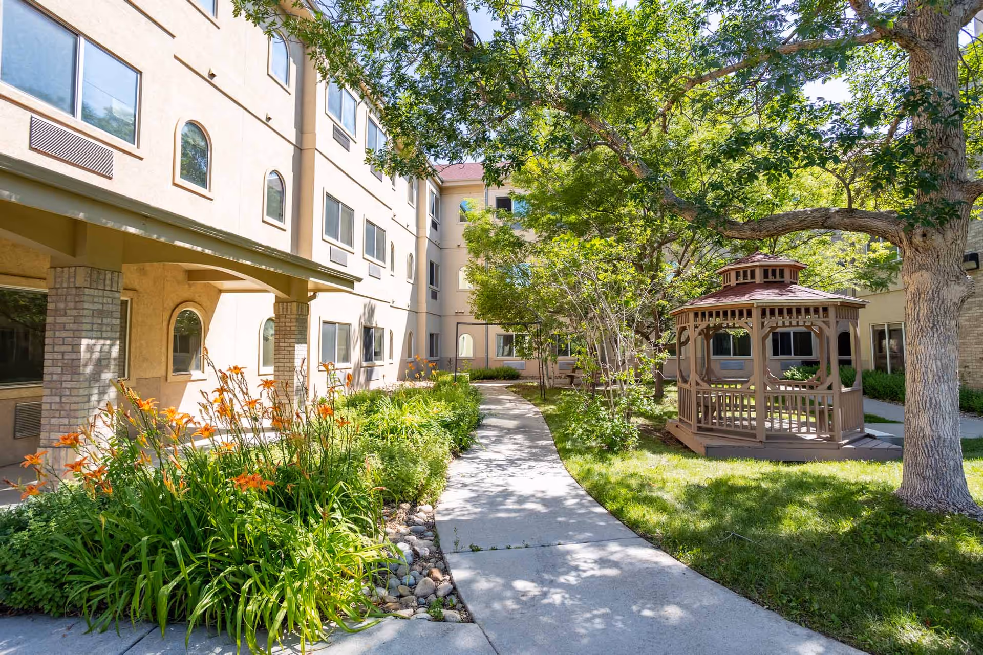 Outdoor courtyard area of Marycrest Assisted Living featuring a paved walkway, green grass, blooming orange flowers, trees providing shade, and a wooden gazebo near the building.