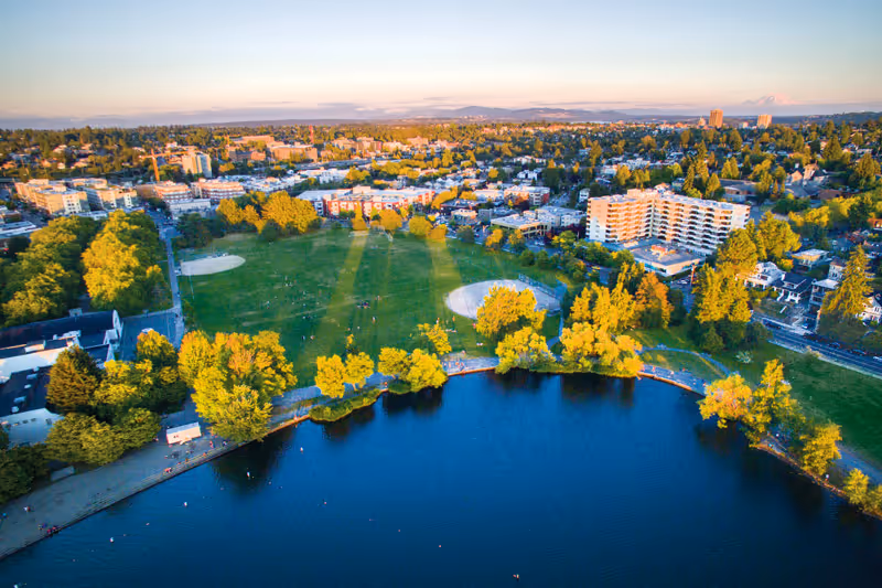 Aerial view of a lakeside park with grassy fields, trees, a shoreline and nearby apartment buildings.
