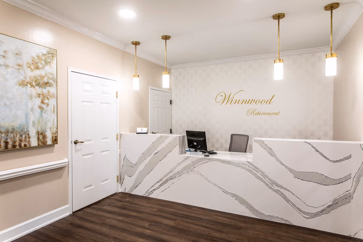 Reception desk in a retirement community lobby with a marble-patterned counter, pendant lights, and a 'Winnwood Retirement' sign on the back wall.