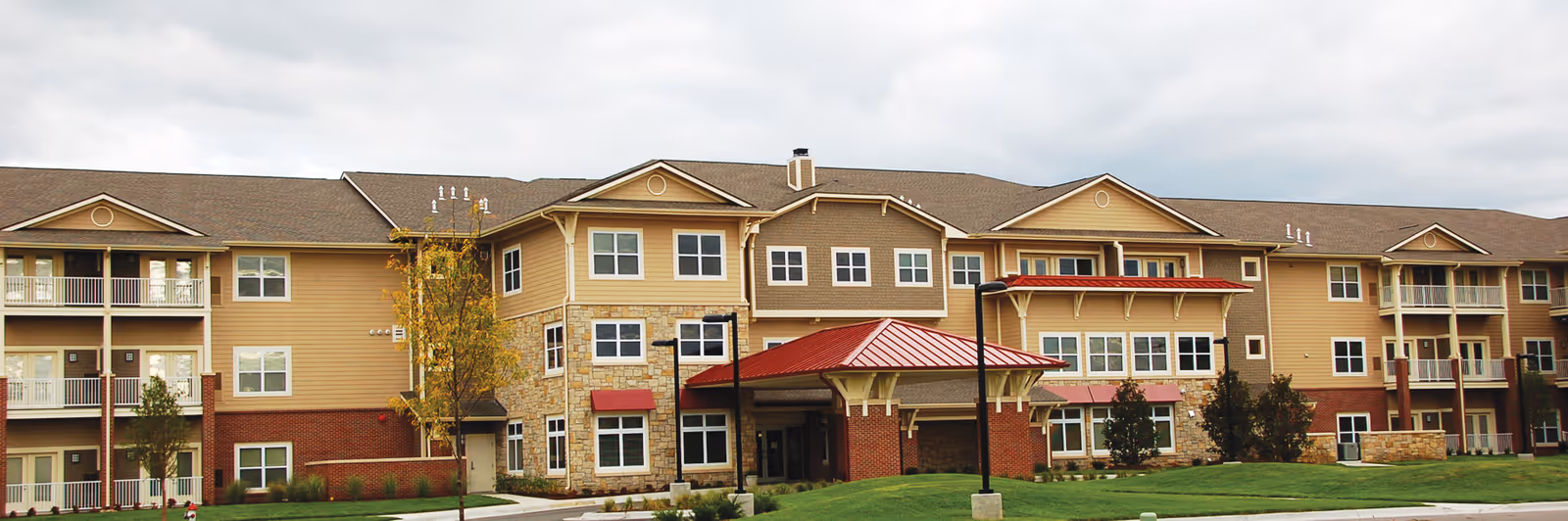 Front exterior of a multi-story senior living building with balconies, stone and brick siding and a covered red-roofed entrance.