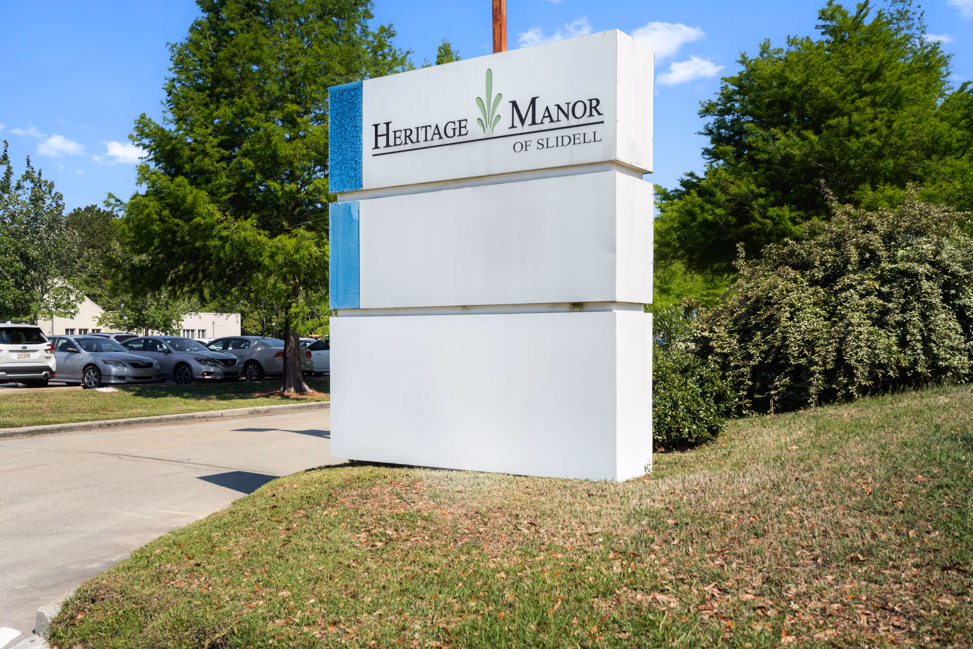 Large white roadside sign reading "Heritage Manor of Slidell" beside a driveway, parked cars, and landscaped greenery.