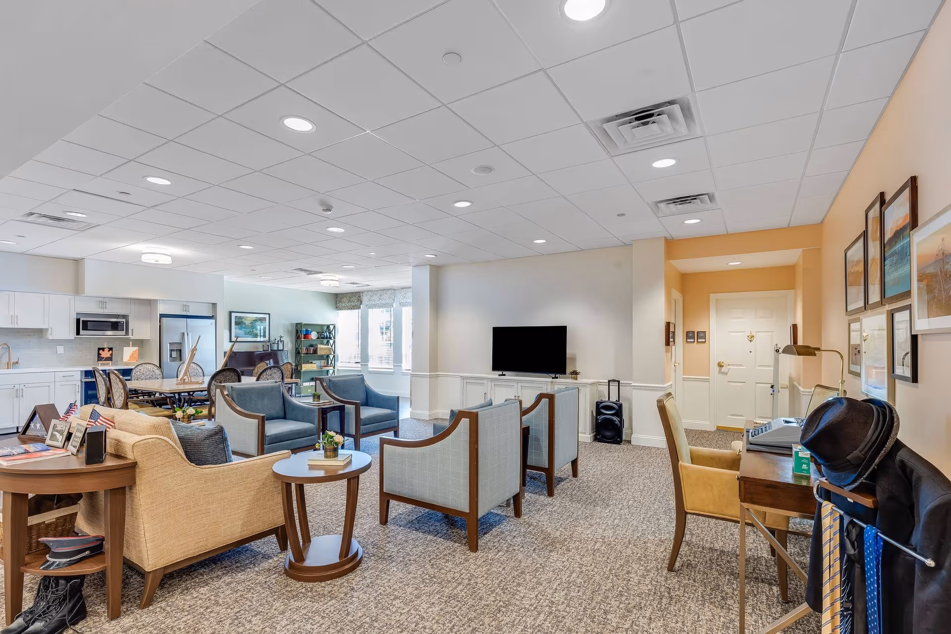 A spacious and well-lit common area in a senior living facility featuring a seating arrangement with armchairs and a sofa around a coffee table, a flat-screen TV mounted on a cabinet, a dining table with chairs, and a kitchenette with white cabinets and stainless steel appliances. The room has carpeted floors, a drop ceiling with recessed lighting, and walls decorated with framed artwork. There is also a desk with a chair and a coat rack holding a jacket, hat, and ties.