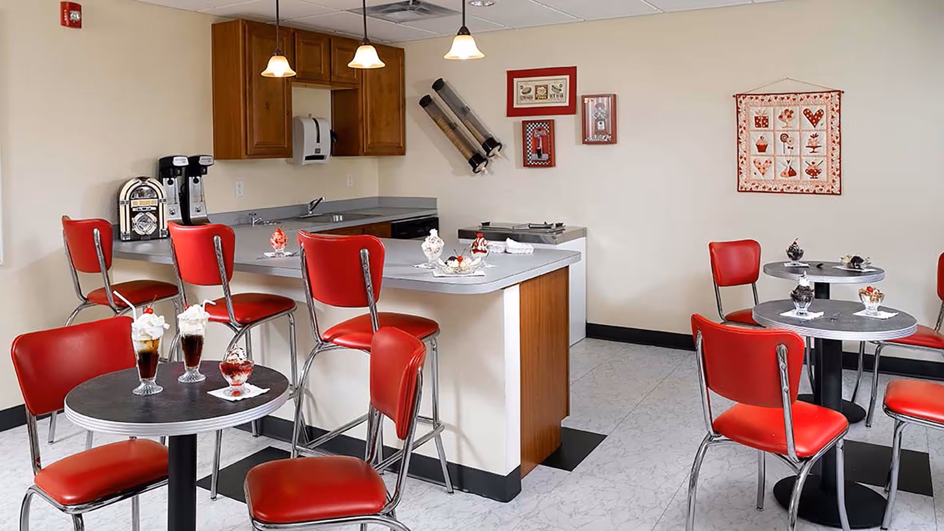 A retro-style dining area with red cushioned chairs and round black tables. The room features a counter with bar stools, a small kitchen area with wooden cabinets, and various framed decorations on the beige walls. Several dessert dishes are placed on the tables and counter.